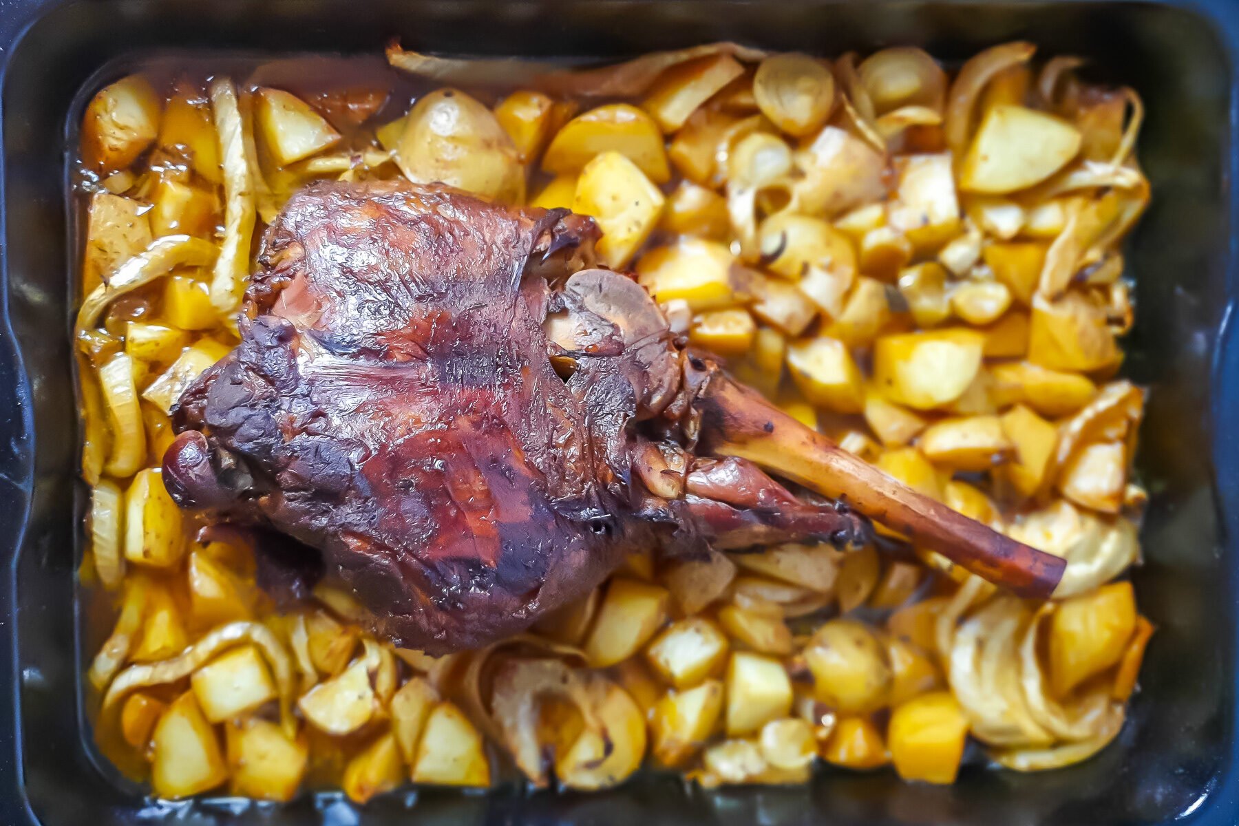 Greek leg of lamb roast on a bed of potatoes and fennel, in a black baking dish.