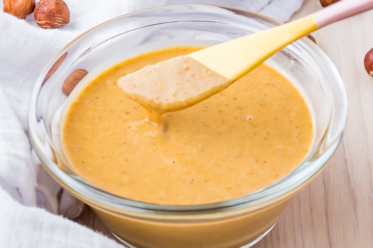 A bowl of hazelnut butter with a spatula rests on a white cloth, surrounded by scattered whole hazelnuts.