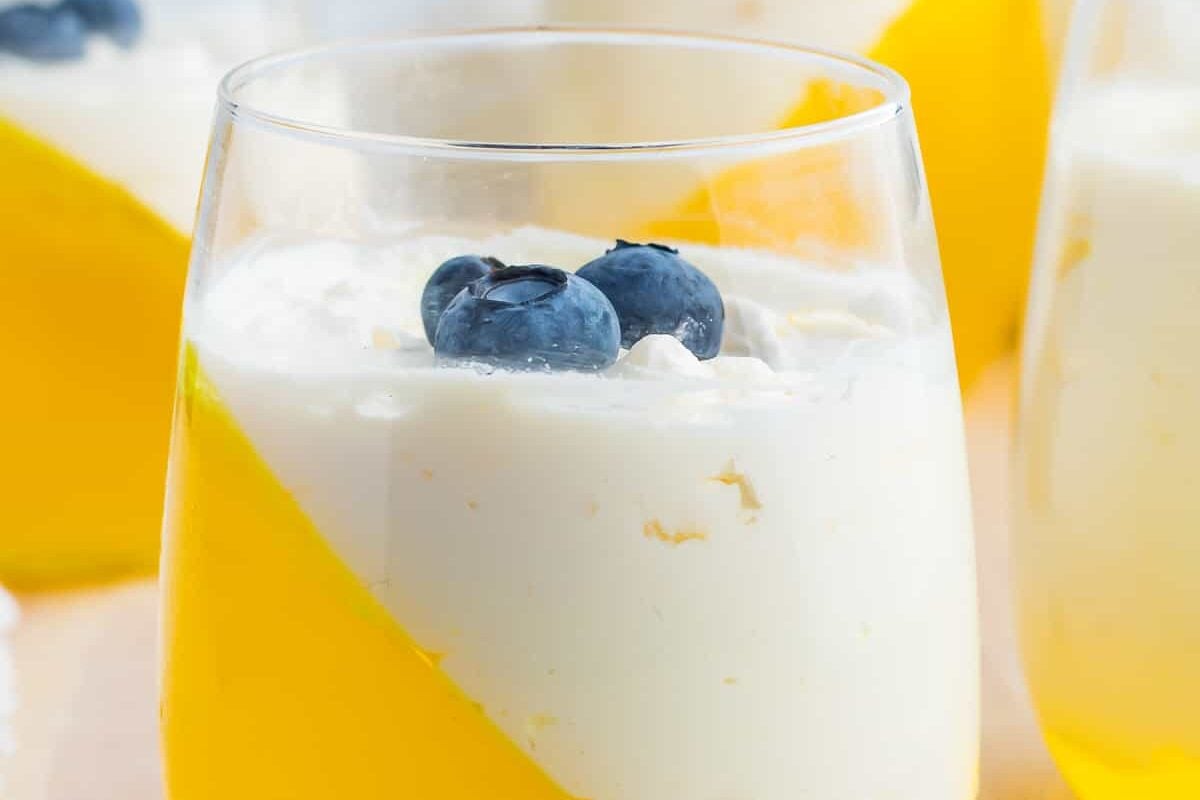 Yellow jello with whipped cream in a stemless wine glass garnished with blueberries.