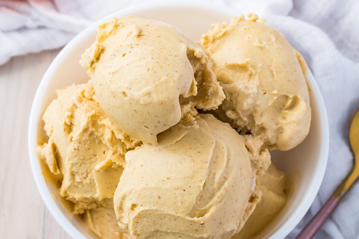 A bowl of creamy, keto pumpkin ice cream scoops, surrounded by a white cloth. A small pumpkin is partially visible in the background, and a gold spoon rests nearby.