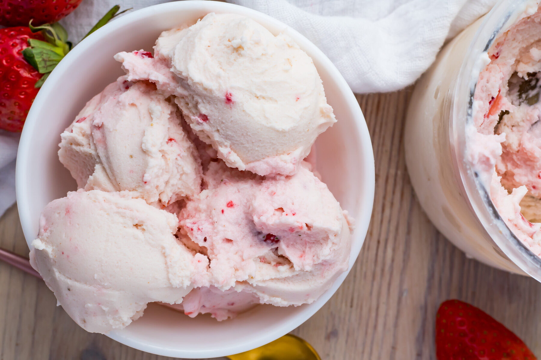 A bowl of creamy ninja creami keto strawberry ice cream with visible strawberry chunks. The table also has fresh strawberries, a spoon, and a white cloth nearby. A partially eaten ice cream container is in the background.