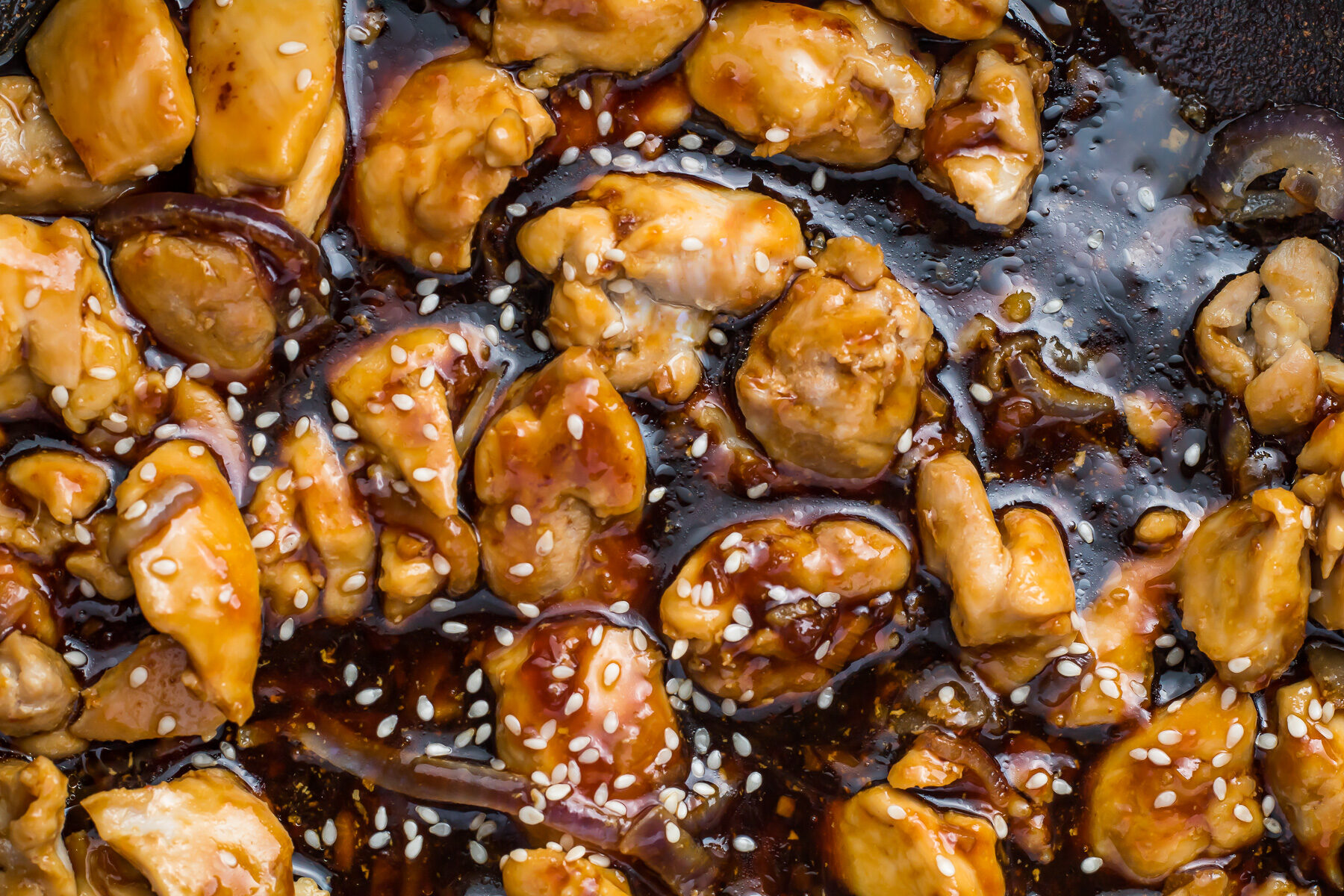 Close-up of cooked chicken pieces coated in a sticky teriyaki glaze, sprinkled with sesame seeds. The chicken is golden brown and shiny, highlighting the rich sauce.