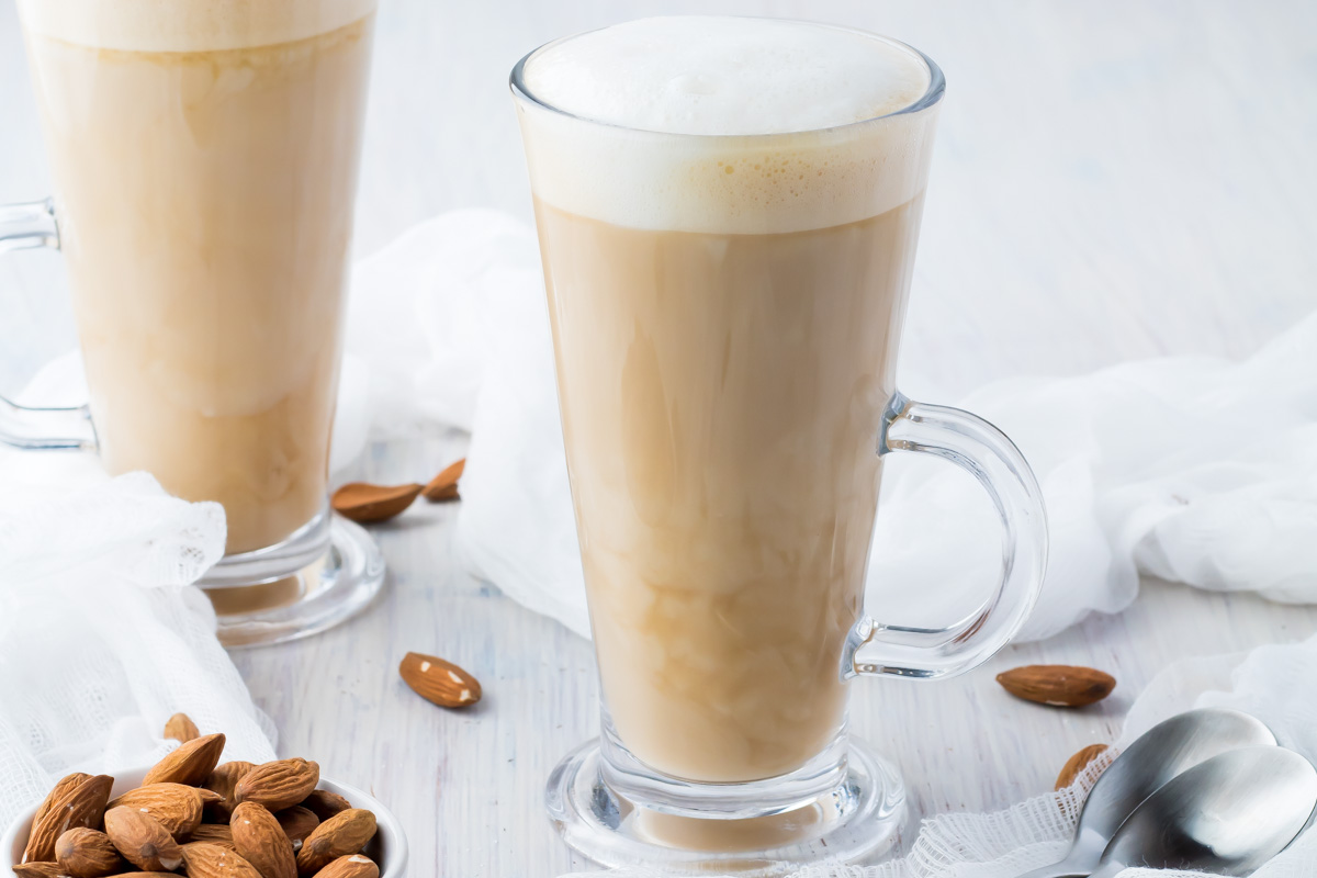A glass mug filled with frothy almond milk latte sits on a light surface, surrounded by whole almonds and a spoon. Another latte is partially visible in the background.