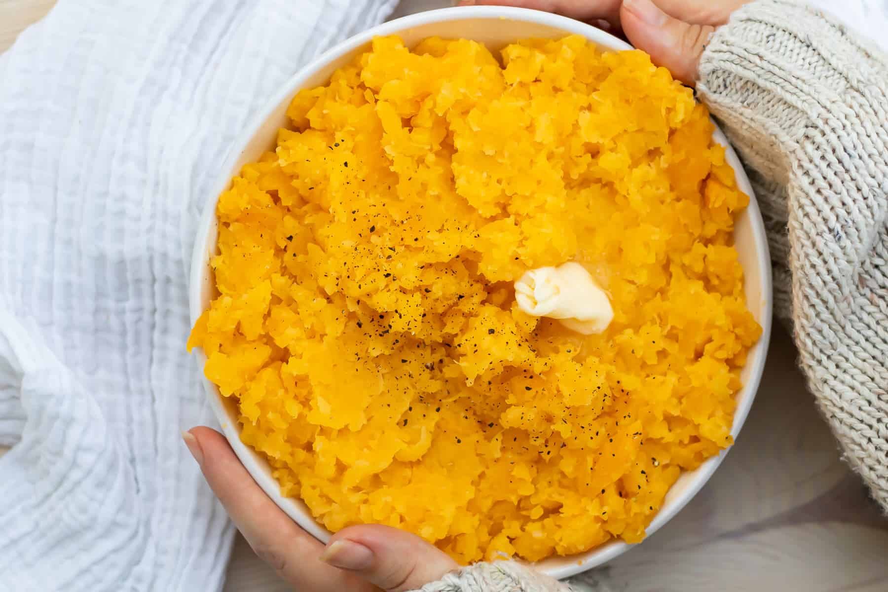 Overhead picture of hands holding a bowl of mashed rutabaga next to a white cloth.