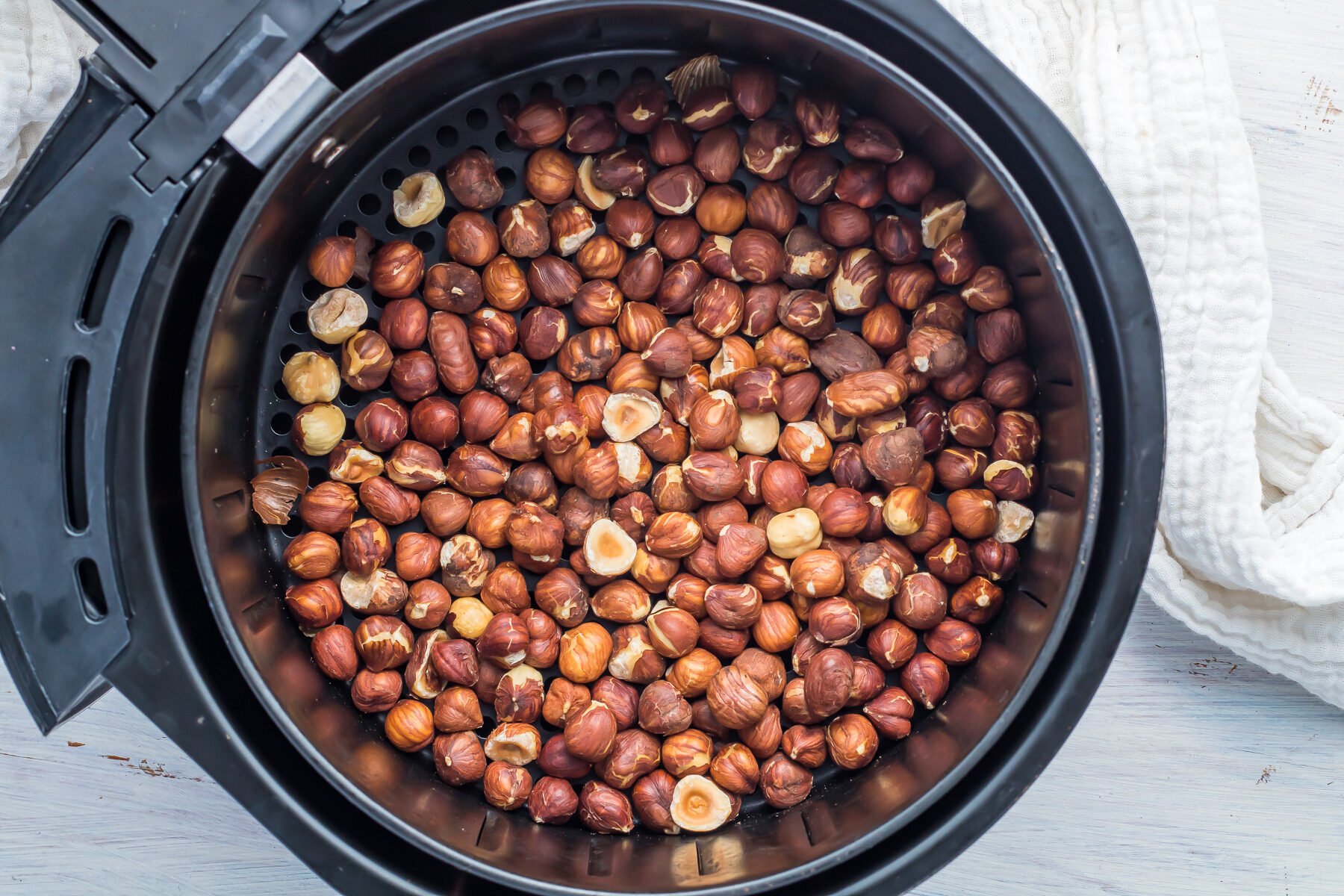 A black air fryer basket filled with roasted hazelnuts. The nuts have a rich brown color, and some shells are cracked. A textured white cloth is partially visible in the background on a light surface.
