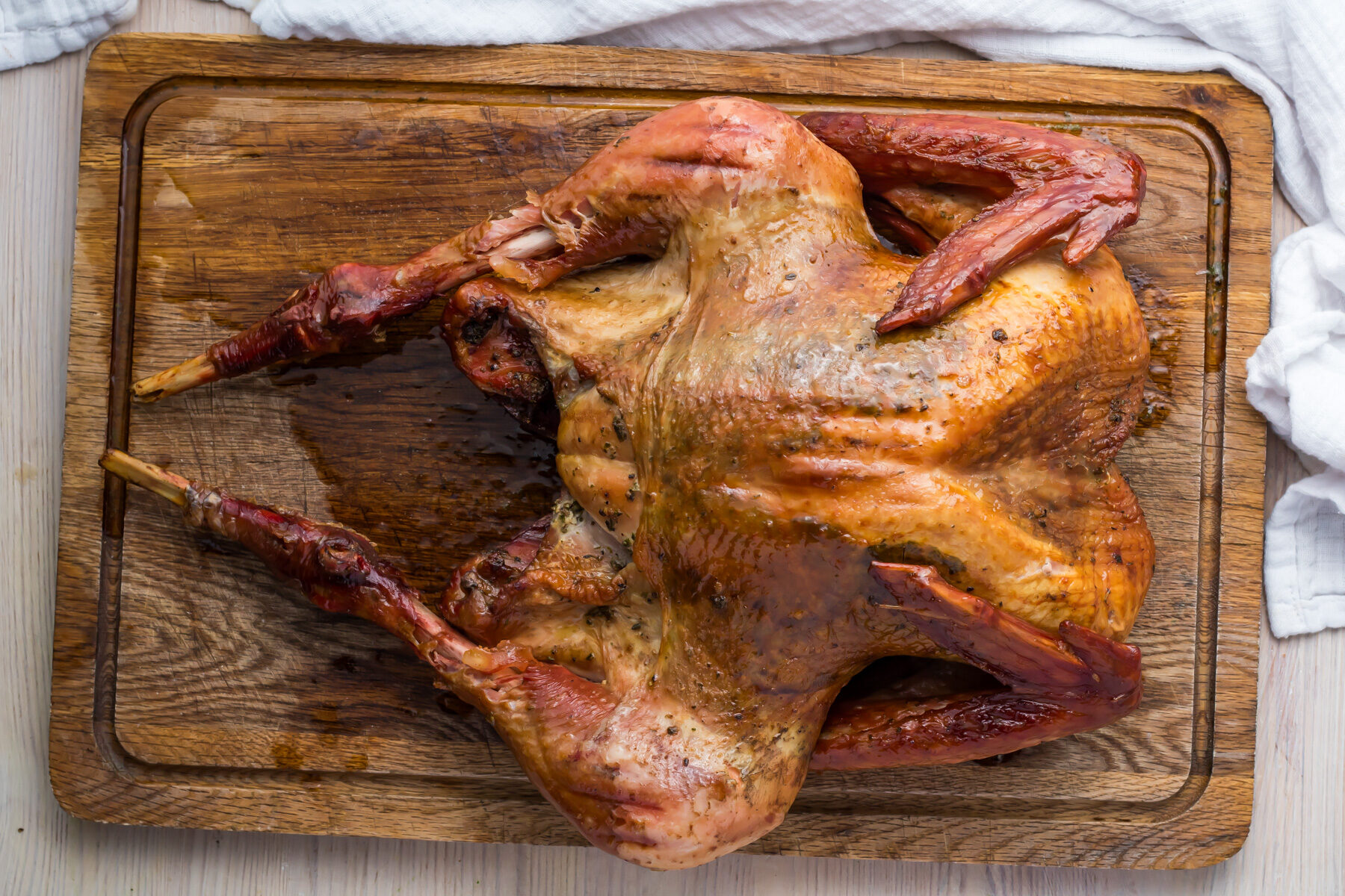 Smoked turkey spatchcocked on a wooden cutting board, cooked to a golden brown color, with crispy skin. A white cloth is partially visible in the top left corner.