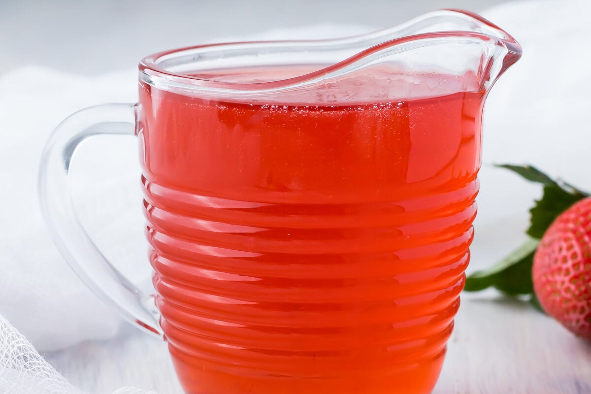 A jug of strawberry syrup on a table with strawberries and a white cloth.