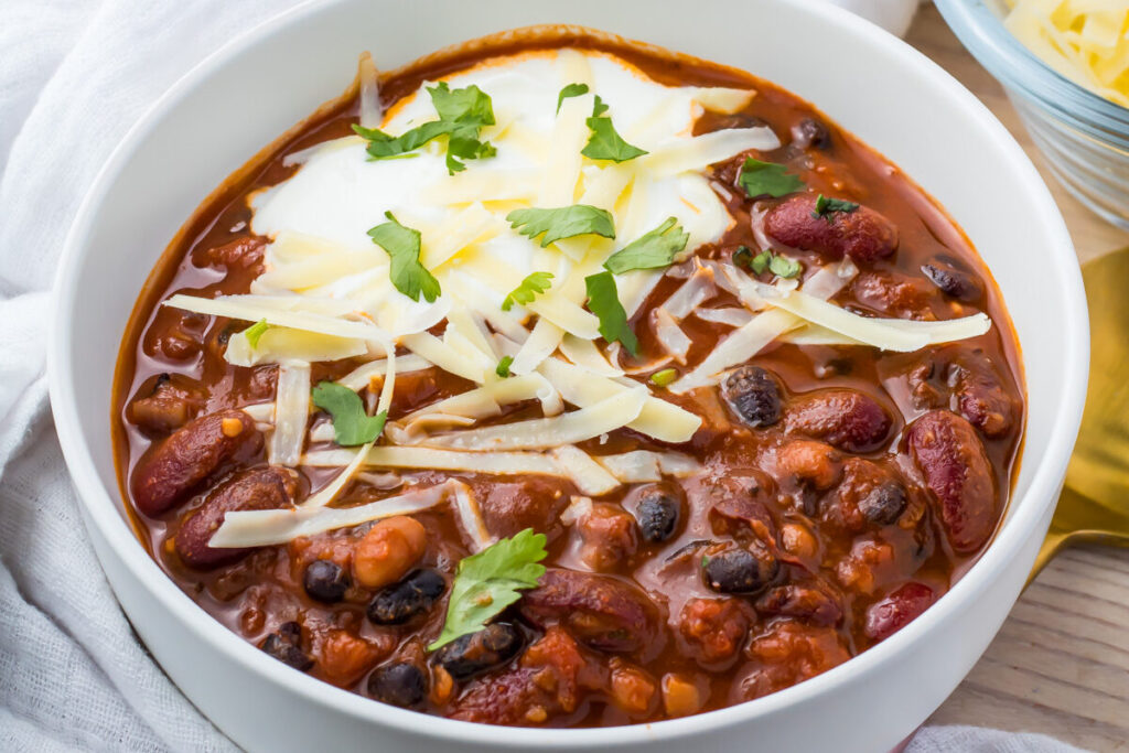 Two bowls of vegan three bean chili topped with shredded cheese and cilantro sit on a white cloth. A dollop of sour cream garnishes each serving. A small bowl of additional cheese and part of a pressure cooker are visible in the background.