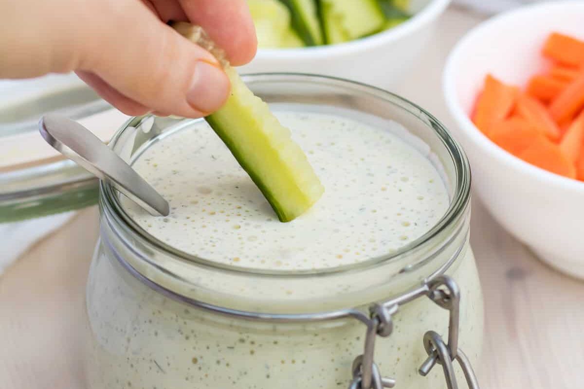 A person is dipping cucumbers into a jar of ranch dressing.