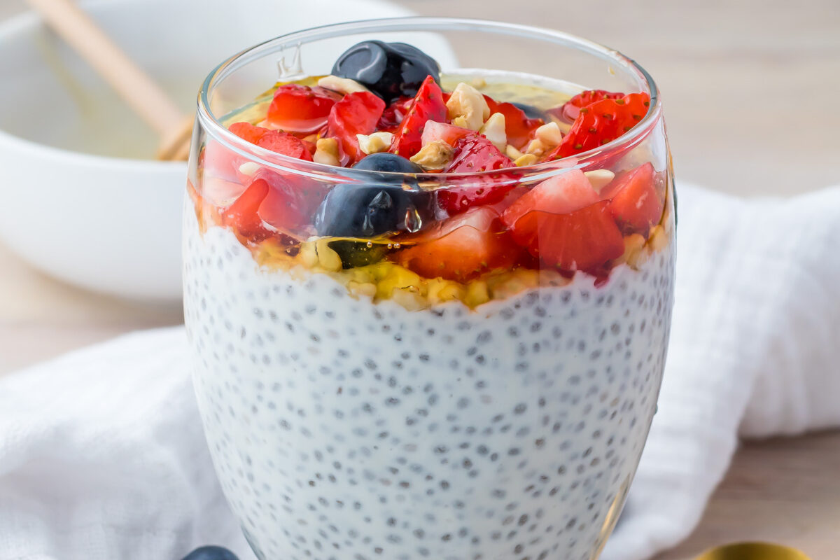 A glass of yogurt chia pudding topped with diced strawberries, blueberries, chopped nuts, and honey sits on a table beside a gold spoon and scattered blueberries.