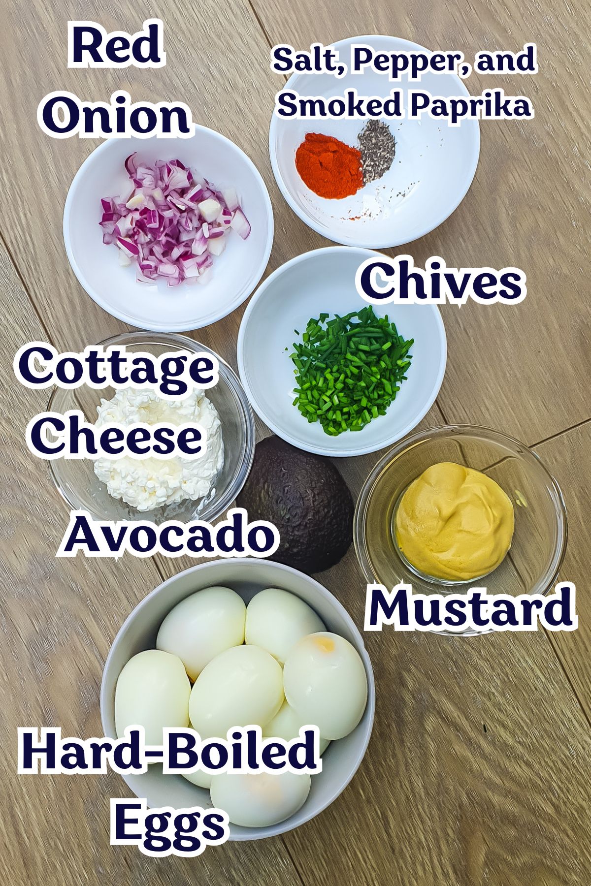 A wooden surface with bowls containing chopped red onion, chives, cottage cheese, mustard, hard-boiled eggs, an avocado, and a bowl of salt, pepper, and smoked paprika.