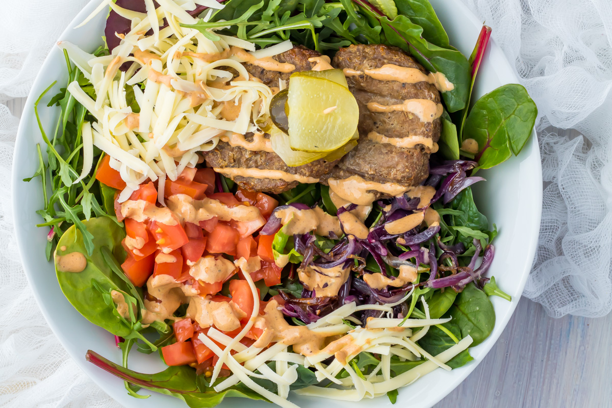 A cheeseburger bowl of salad with leafy greens, chopped tomatoes, shredded cheese, grilled patties, lemon wedge, and creamy dressing drizzled on top.