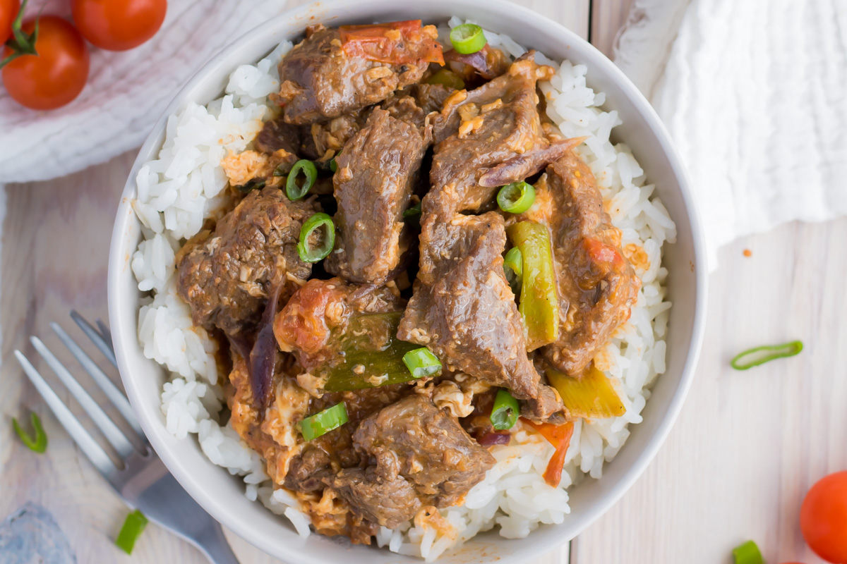 A bowl of white rice topped with beef stew, green onions, and vegetables, placed on a light wooden surface with a fork and tomatoes nearby.