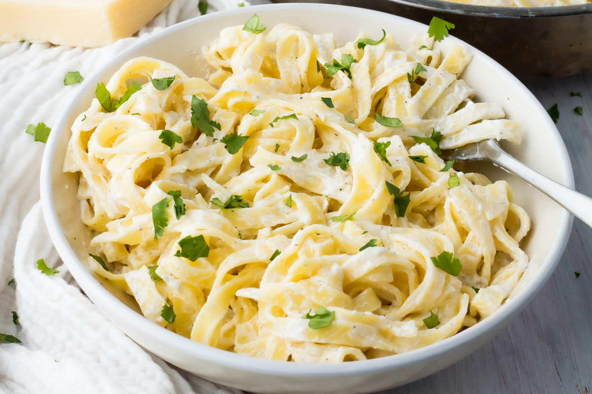 A bowl of cottage cheese fettuccine Alfredo garnished with chopped parsley, with a fork resting on the side.