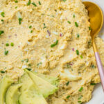 A bowl of cottage cheese avocado egg salad garnished with sliced avocado and chopped chives, with a gold spoon resting on the side.