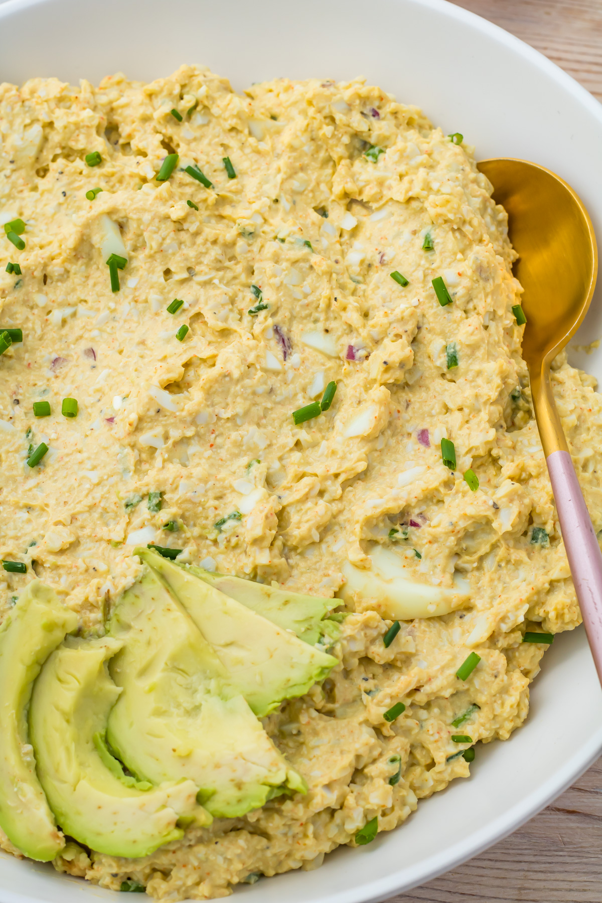 A bowl of cottage cheese avocado egg salad garnished with sliced avocado and chopped chives, with a gold spoon resting on the side.