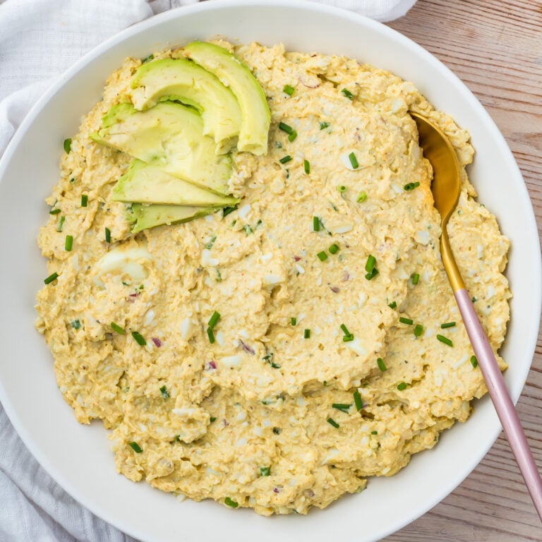 A bowl of creamy cottage cheese avocado egg salad topped with sliced avocado and chopped chives, served with a gold spoon on the side.