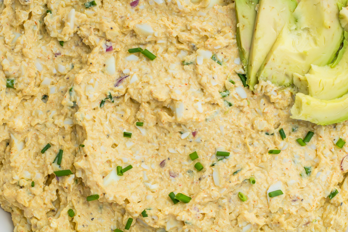 Close-up of a creamy cottage cheese avocado egg salad mixed with chopped chives and spices, topped with sliced avocado on the side.