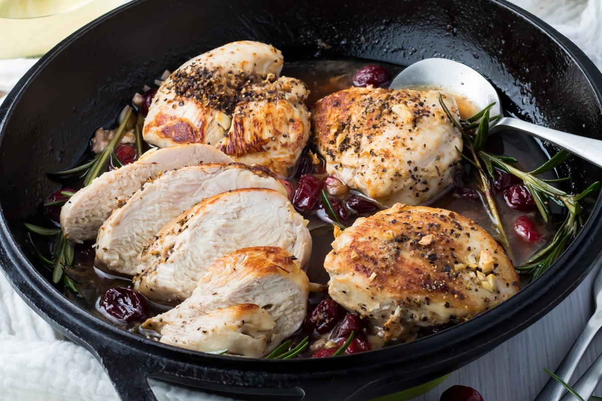 Sliced, seasoned chicken breast in a cast-iron skillet with cranberries, fresh rosemary, and a light sauce&mdash;served with a metal spoon.