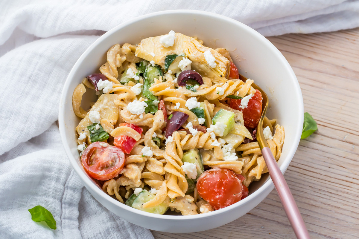 A bowl of pasta salad with cherry tomatoes, cucumber, olives, spinach, artichoke, and crumbled feta cheese, with a fork resting on the bowl.