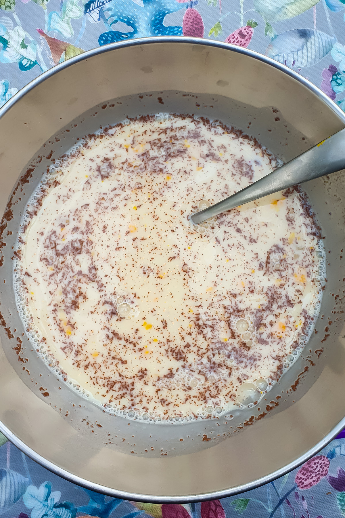 A metal bowl containing a mixture of milk, eggs, and grated chocolate with a metal spoon in it, on a floral tablecloth.
