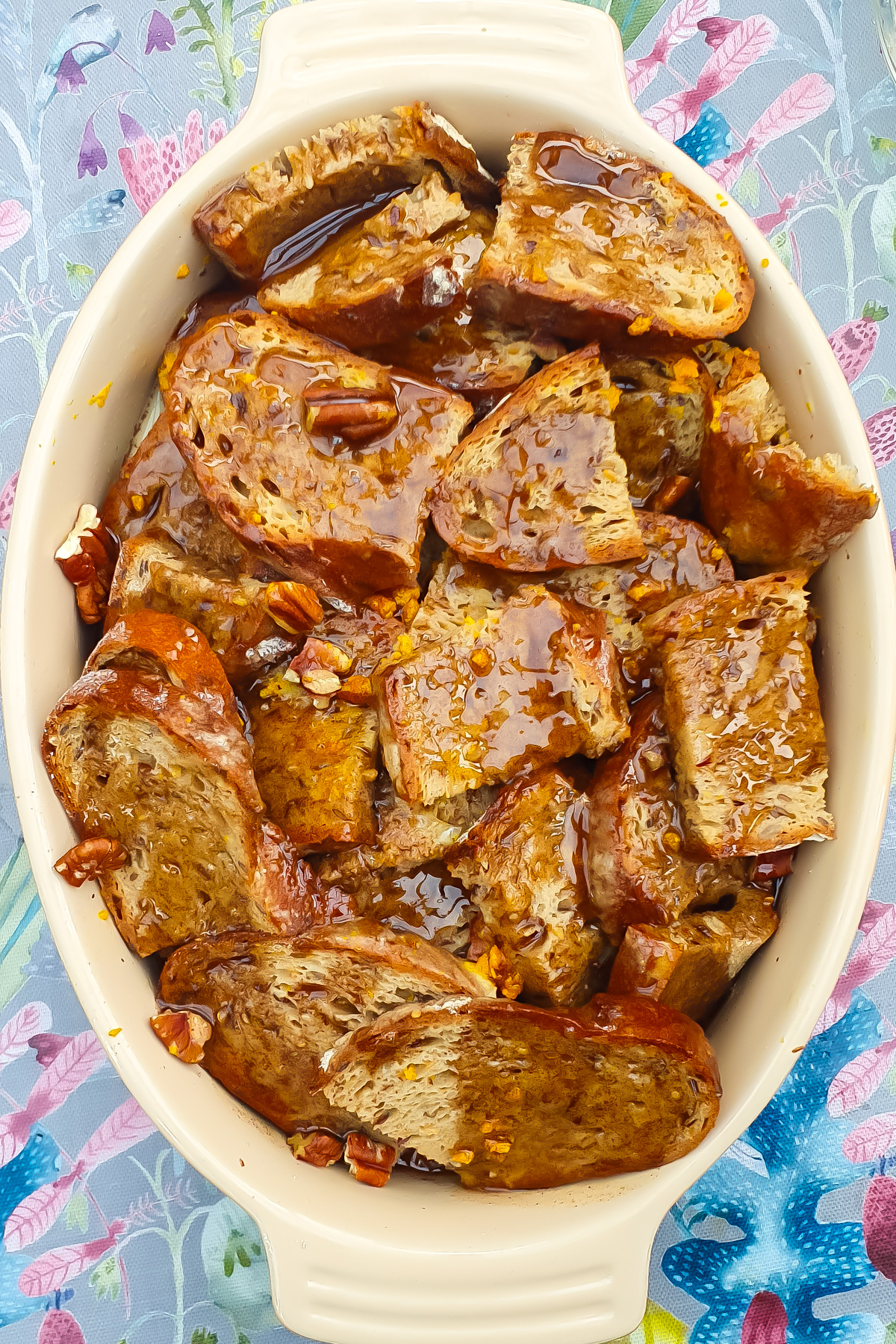 Sliced bread soaked in a brown syrupy mixture with chopped nuts, arranged in a white oval baking dish on a patterned tablecloth.