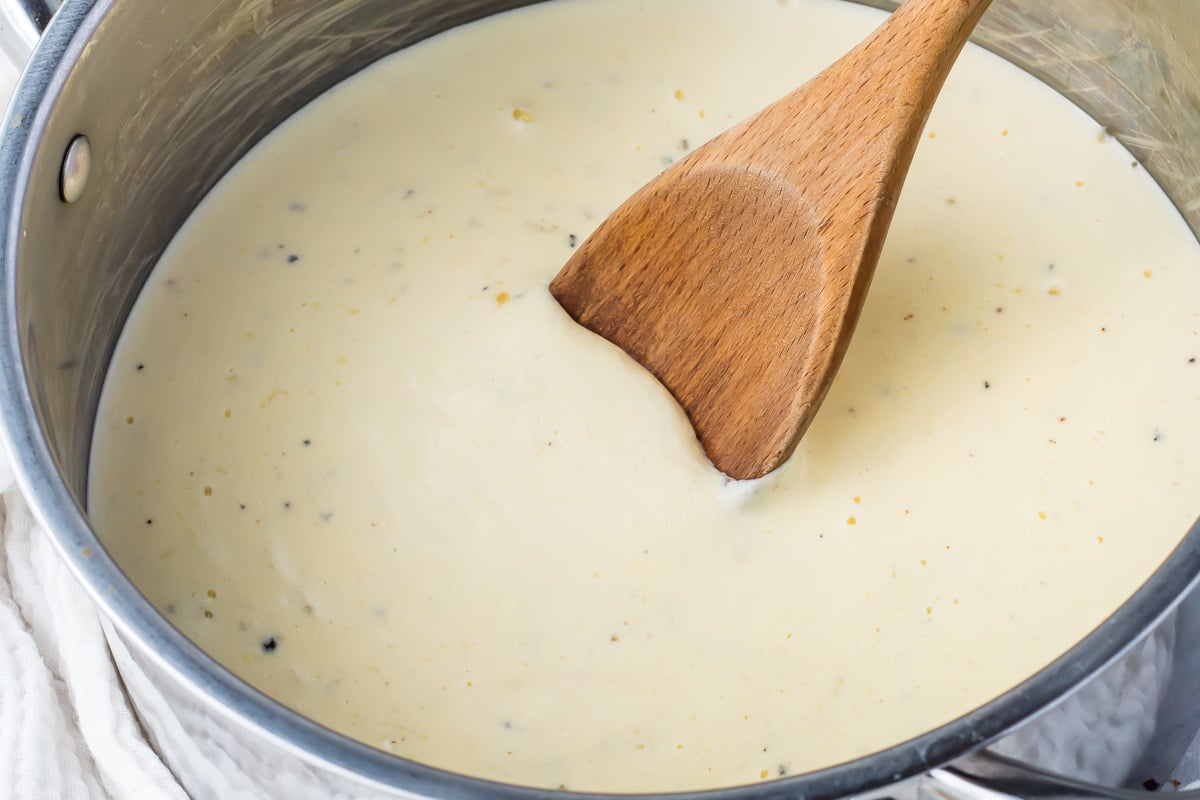 A wooden spoon stirs a creamy white keto alfredo sauce with black pepper specks in a stainless steel pot.