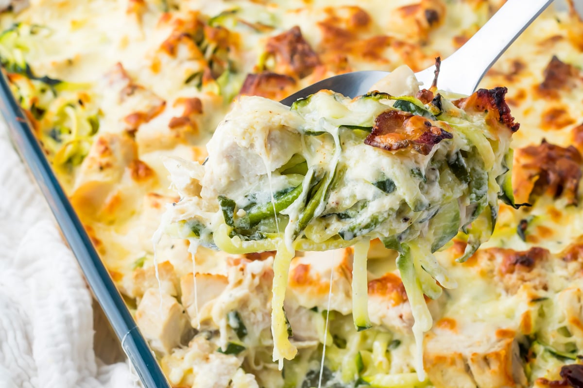 A close-up of a baked chicken alfredo casserole with cheese, chicken, and vegetables, with a serving being lifted out on a spoon.