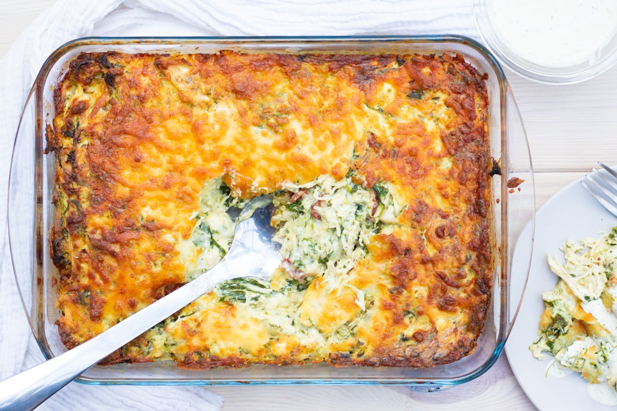A baked casserole with a golden-brown cheesy top, partially served with a large spoon, next to a plate of the casserole and a cup of sauce.