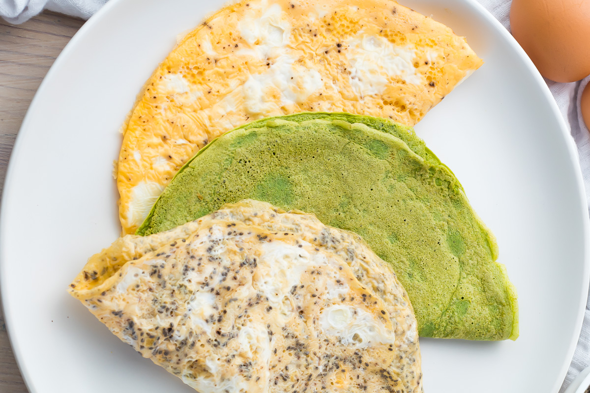 Three folded keto egg wraps—one plain, one green, and one speckled with seeds—are arranged on a white plate.