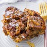 A serving of baked French toast topped with powdered sugar and chopped pecans, with syrup being poured over it, on a white plate with a gold fork.