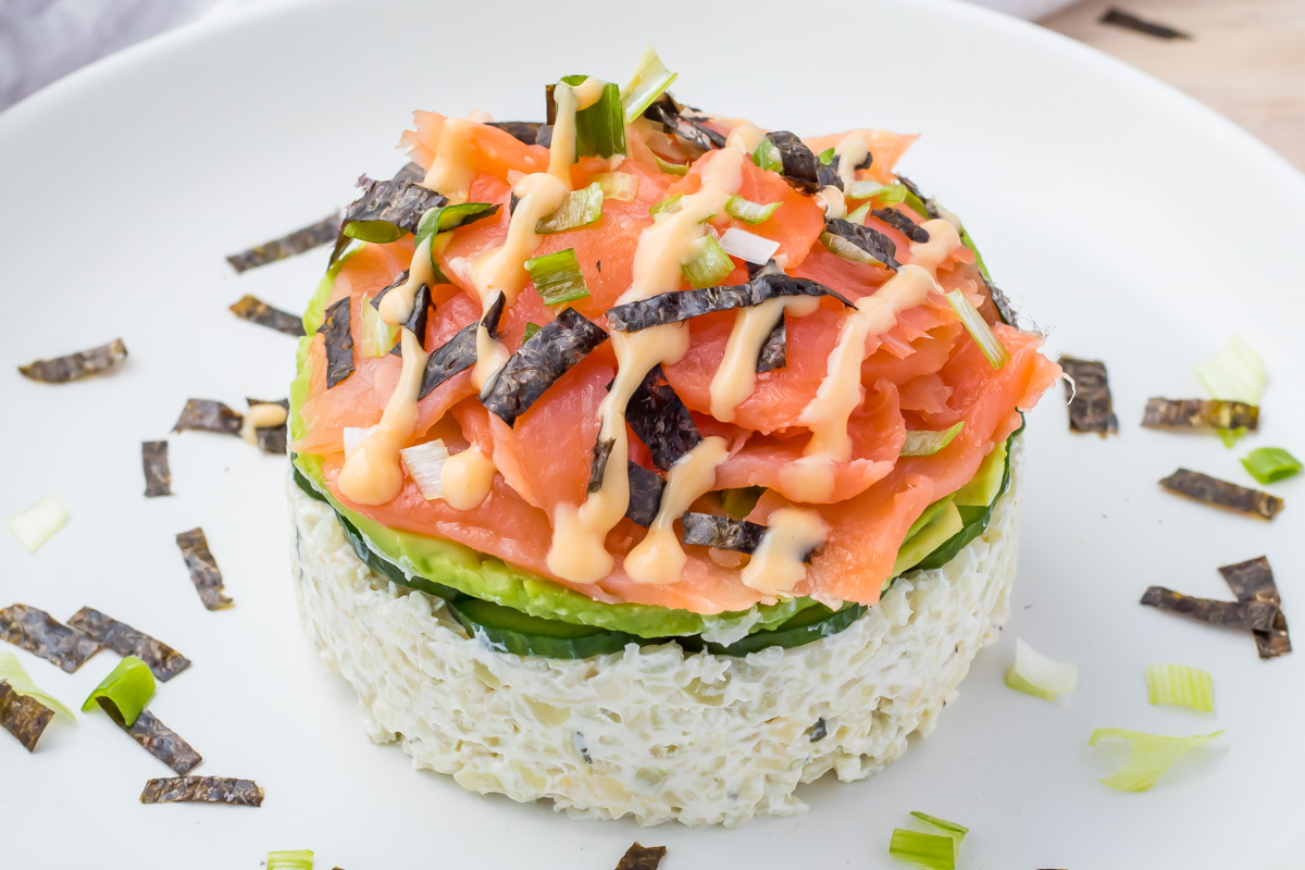 A round serving of sushi-inspired salad topped with sliced salmon, cucumber, seaweed strips, and drizzled sauce on a white plate.