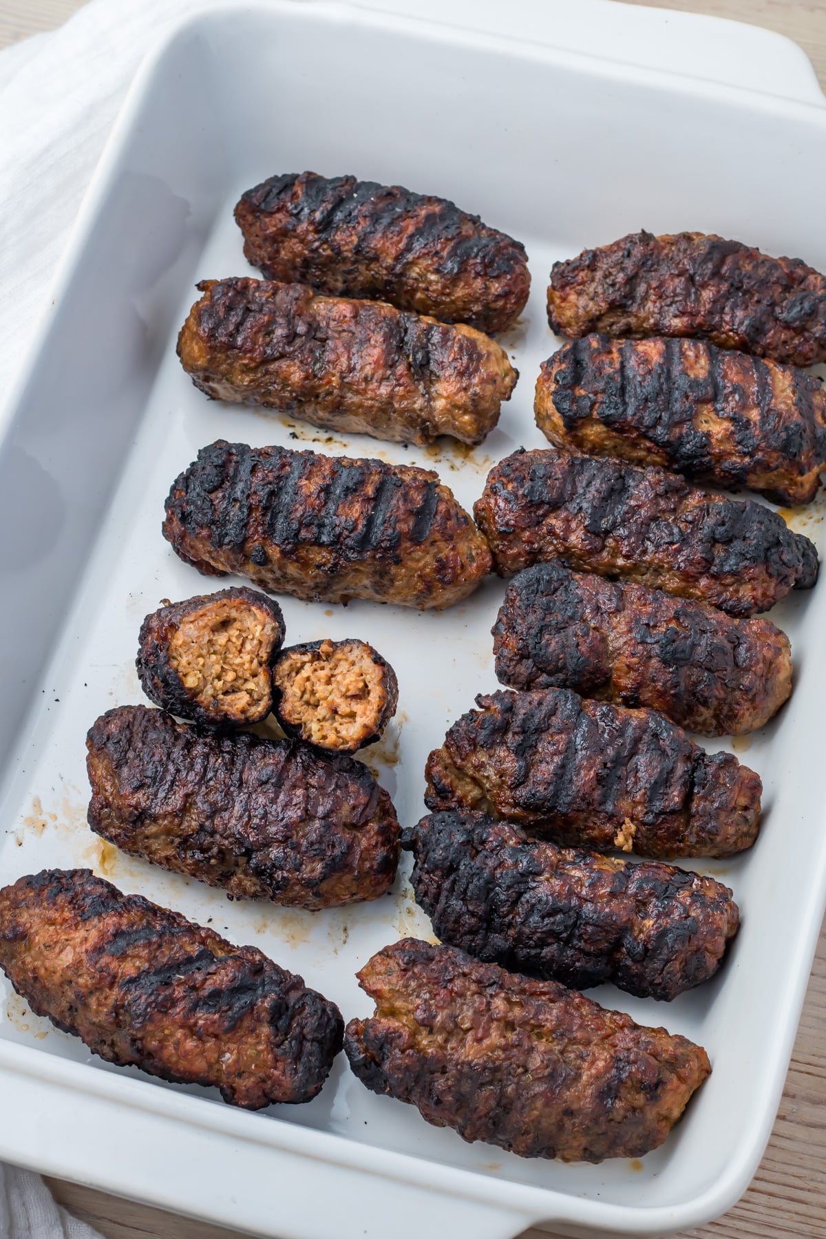 Twelve grilled lamb kofta kebabs are arranged in a white baking dish, with one piece cut open to show the cooked interior.