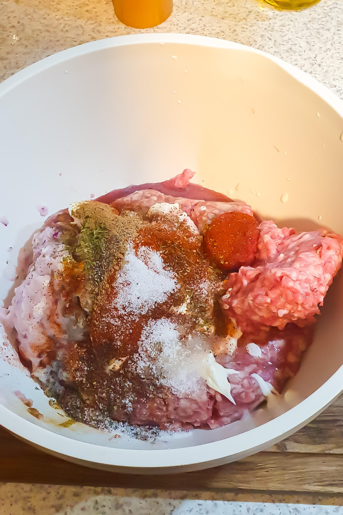 Ground meat with various spices and seasonings on top in a white mixing bowl, placed on a kitchen countertop.