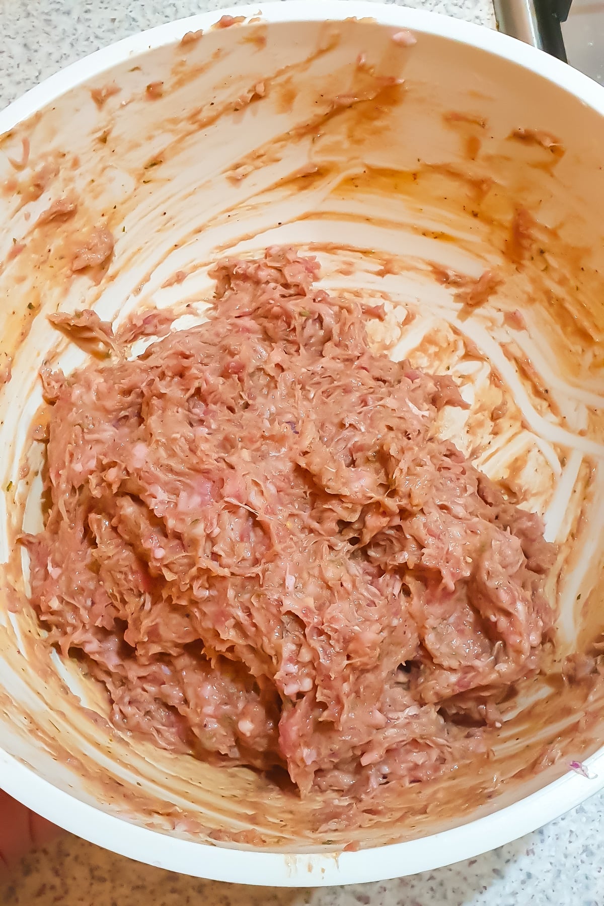 A white bowl containing a mixture of raw ground meat and seasonings, resting on a kitchen countertop.