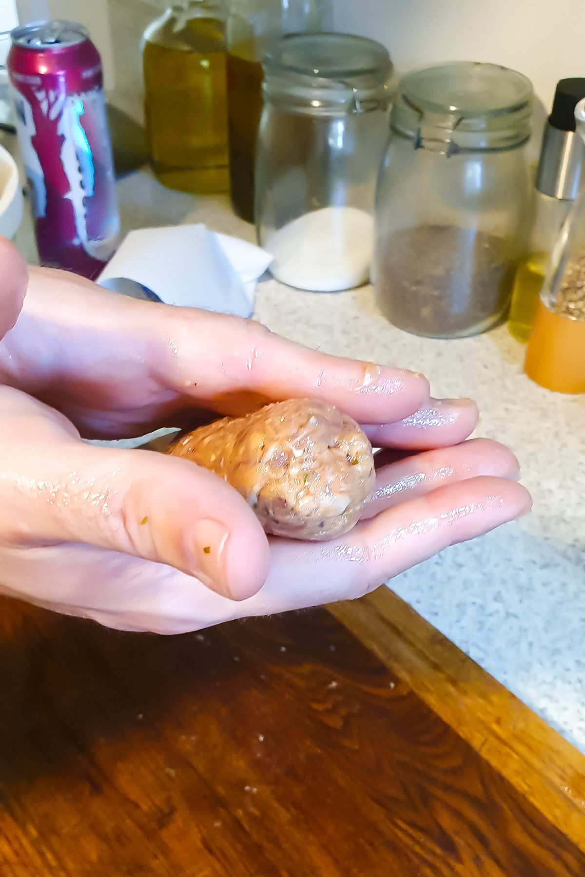 A person shapes a mixture into a log shape with both hands in a kitchen, with jars, a drink can, and spices visible in the background.