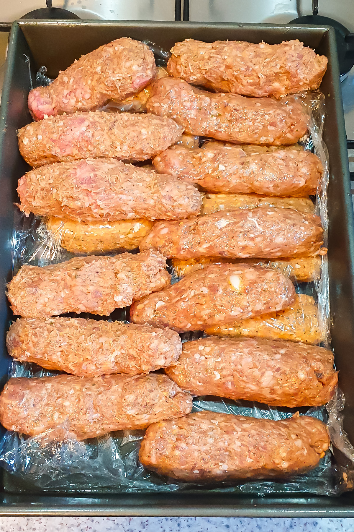 Uncooked lamb kofta kebabs arranged in rows on a tray, each wrapped in plastic wrap, ready for further preparation or cooking.