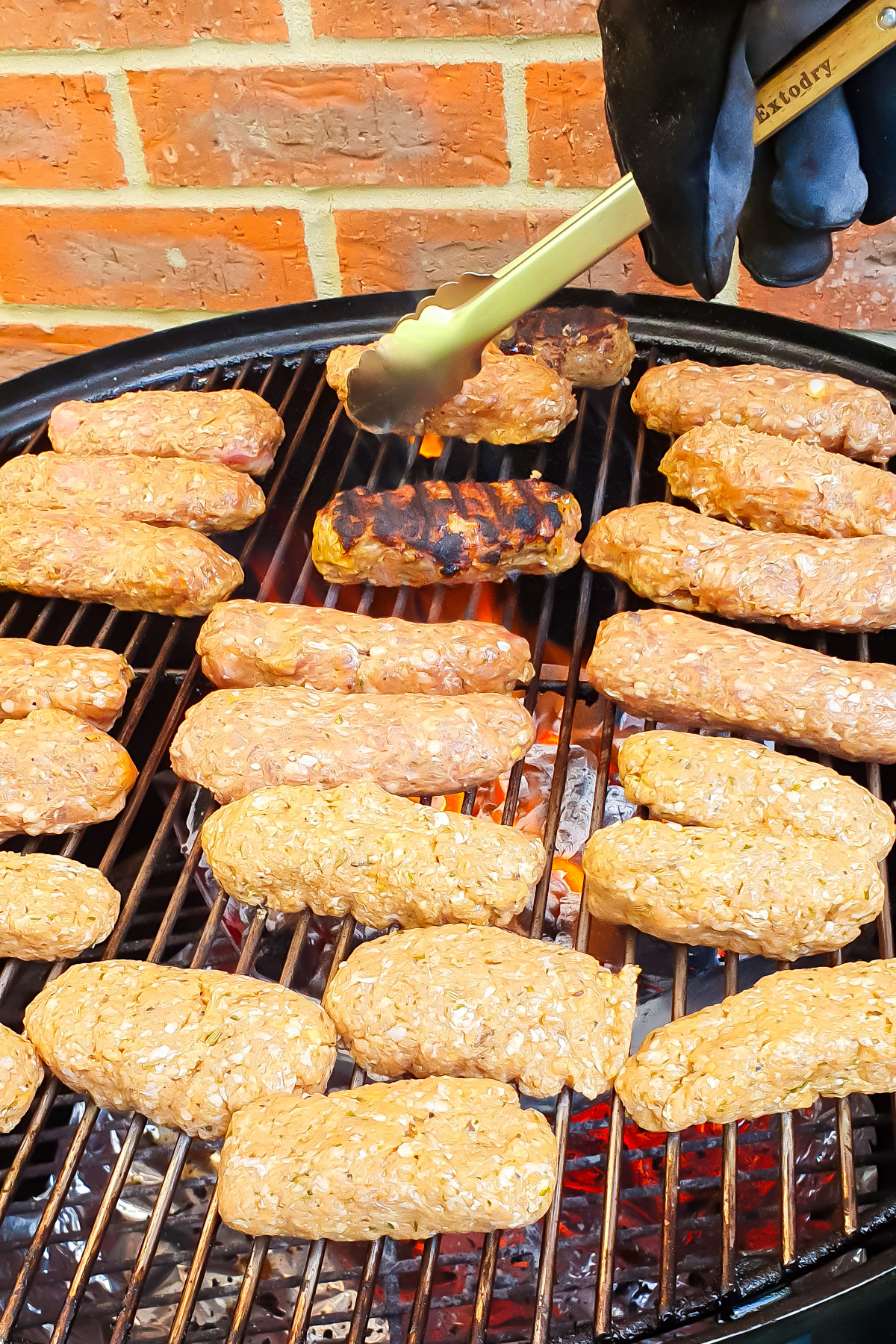 Uncooked and grilled lamb kofta kebabs on a barbecue grill, with a gloved hand using tongs to turn one patty; brick wall in the background.