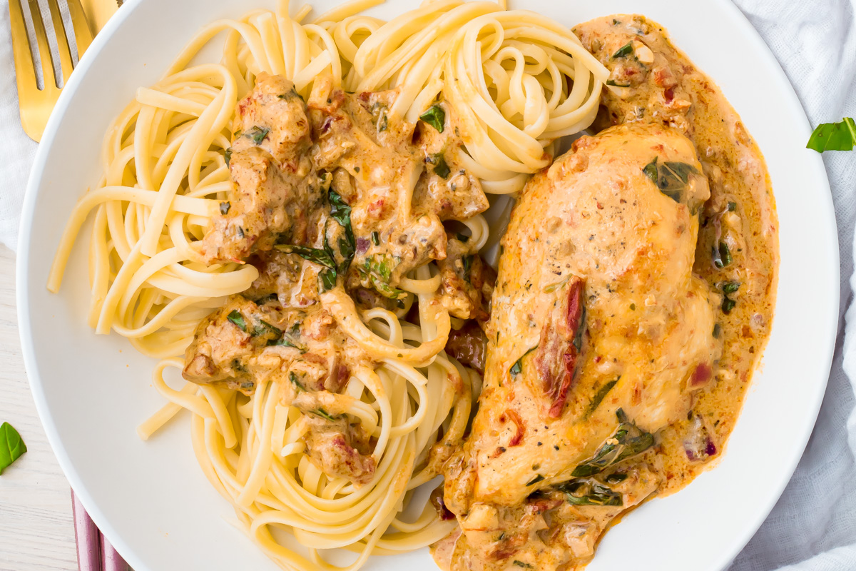 A plate of pasta served with a creamy chicken breast in a herb and tomato sauce, garnished with spinach and sun-dried tomatoes.