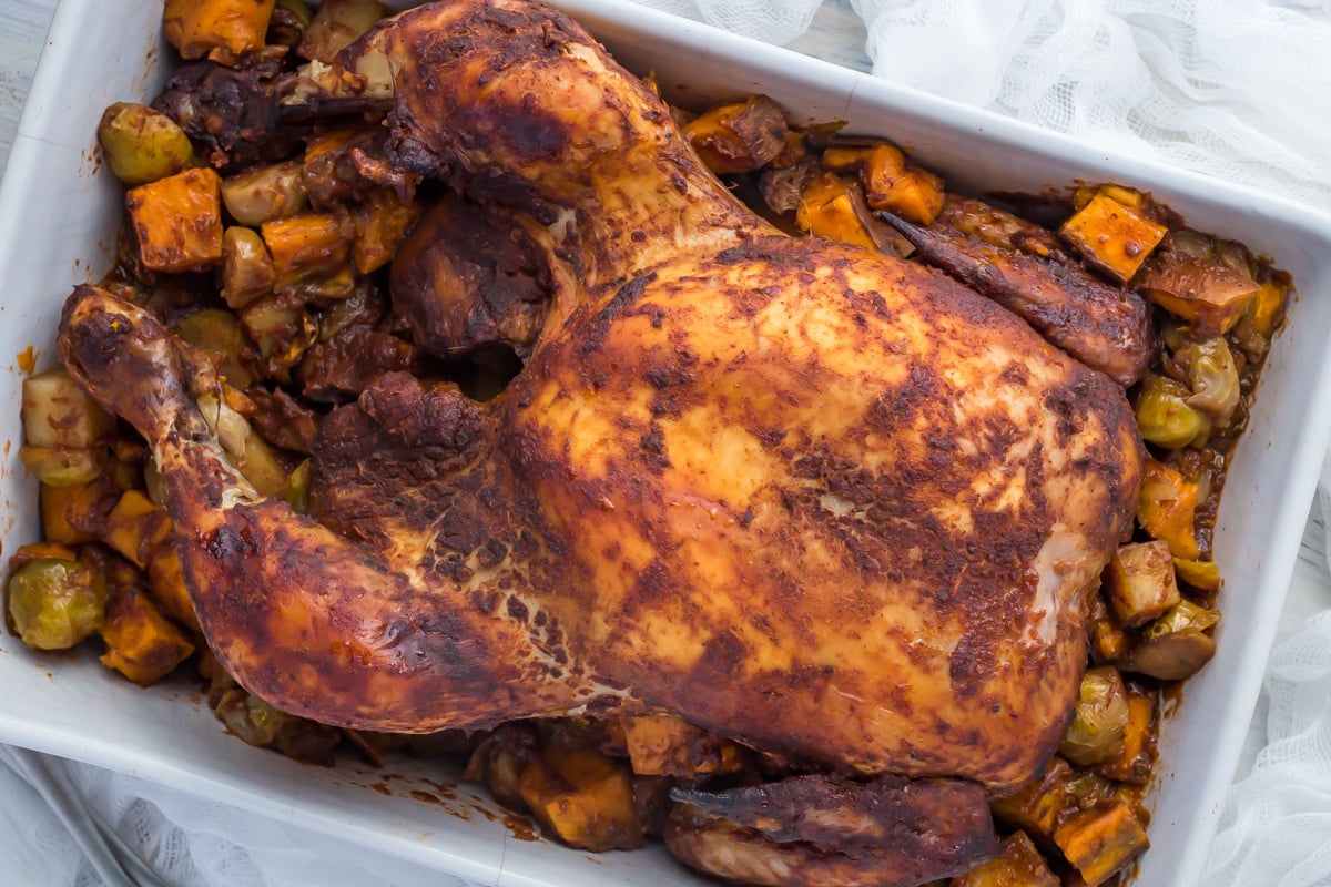 A whole roasted chicken in a baking dish, surrounded by chunks of roasted vegetables like potatoes and Brussels sprouts.