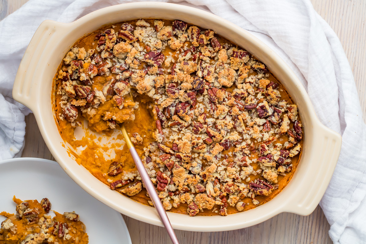 A casserole dish filled with whole30 sweet potato apple casserole topped with pecan crumble, with a serving spoon and a portion served on a plate nearby.