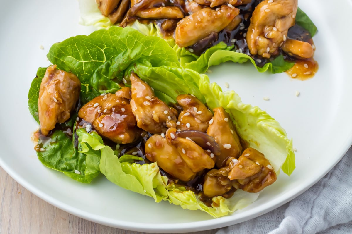Chicken pieces in a glossy sauce served on fresh lettuce leaves, sprinkled with sesame seeds, arranged on a white plate.