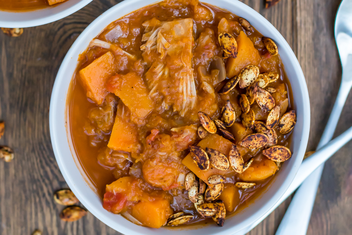 A bowl of thick whole30 vegetable stew with chunks of squash and vegetables, topped with roasted seeds, sits on a wooden surface.