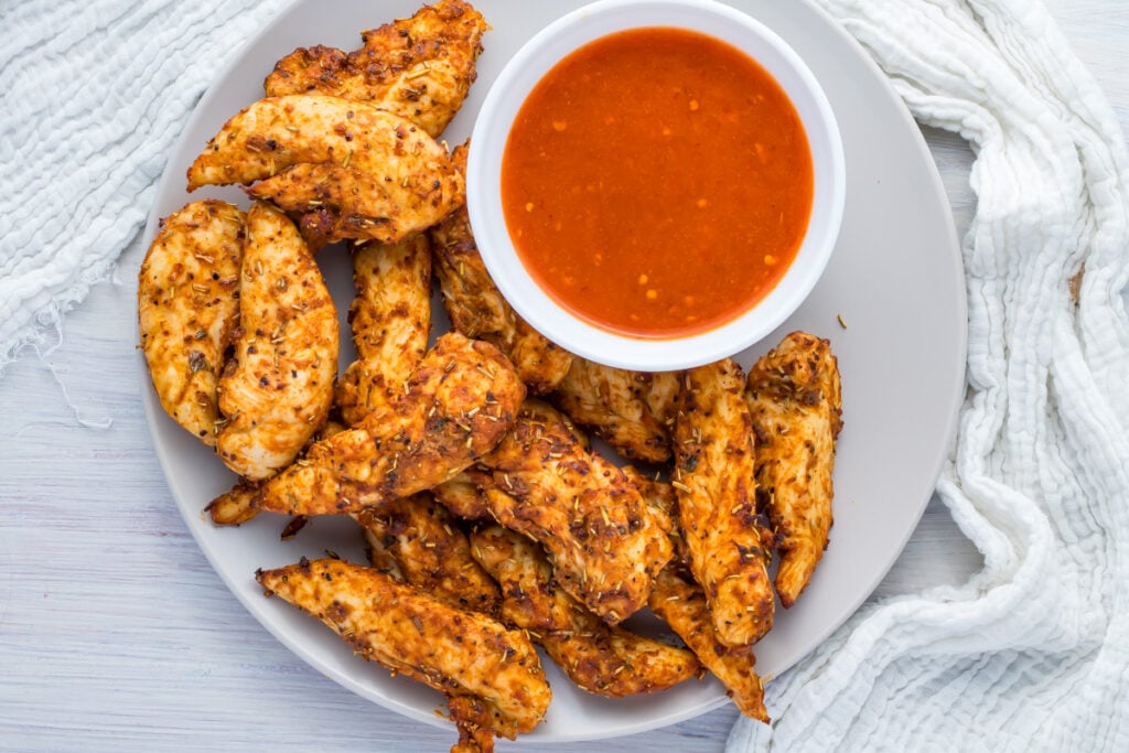 A plate of seasoned whole30 air fryer chicken tenders, arranged around a bowl of red dipping sauce on a light-colored surface.
