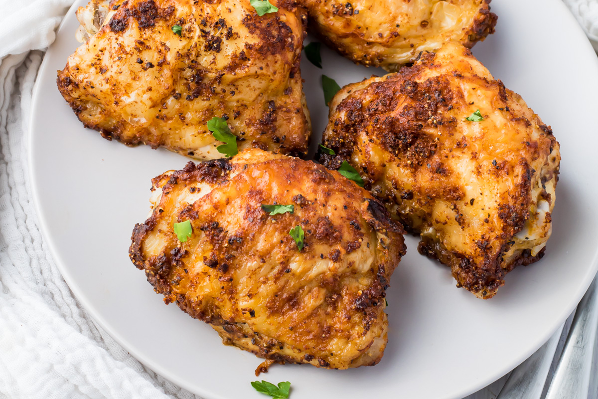Four pieces of seasoned, air fryer whole30 chicken thighs on a white plate, garnished with small parsley leaves.