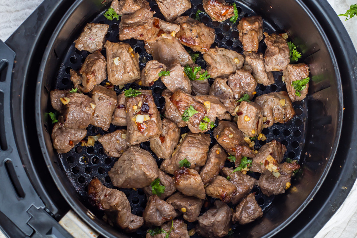Cubed pieces of cooked steak with herbs and garlic inside an air fryer basket.