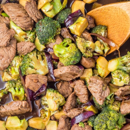A close-up of a whole30 beef and broccoli stir-fry in a pan with a wooden spoon, featuring tender beef, crisp broccoli florets, red onion, and sauce.