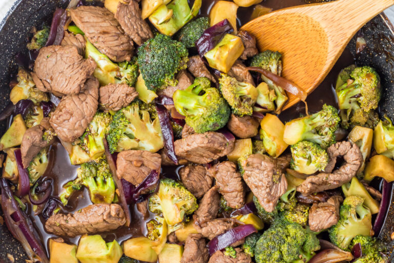 A close-up of a whole30 beef and broccoli stir-fry in a pan with a wooden spoon, featuring tender beef, crisp broccoli florets, red onion, and sauce.