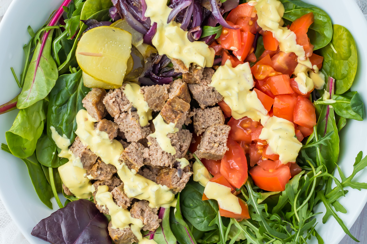 A Whole30 burger salad bowl featuring ground meat, diced tomatoes, pickles, leafy greens, saut&eacute;ed red onions, and a drizzle of yellow dressing on top.