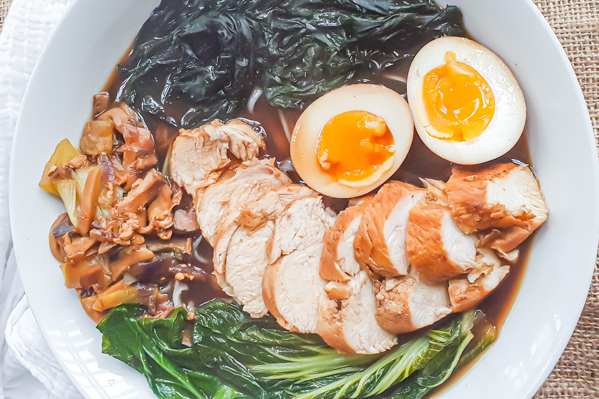 A bowl of whole30 ramen with sliced chicken breast, soft-boiled egg halves, leafy greens, mushrooms, and seaweed in broth.